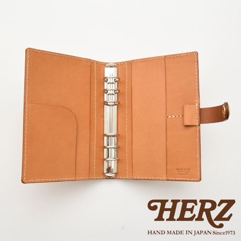 焦糖色皮革手帳，內有六孔活頁夾，左側有卡片插槽，右側有束帶開合，整體以白色縫線勾勒。