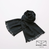 一條深色（接近炭黑色）的今治匹馬棉圍巾，材質輕盈，織紋細緻，兩端帶有流蘇，圍巾的一角捲起，整體呈現簡約優雅的風格。