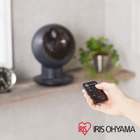 深灰色圓形空氣循環扇，帶有木紋飾面，放置在桌面。一只手正握著黑色遙控器，可見「IRIS OHYAMA」的品牌標誌。