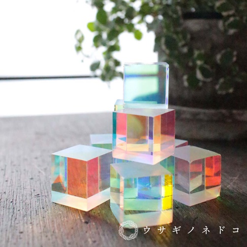 數個透明的稜鏡立方塊堆疊排列，表面呈現彩虹般的光澤，光線穿透時折射出藍、綠、黃、粉等色彩，材質為玻璃。
