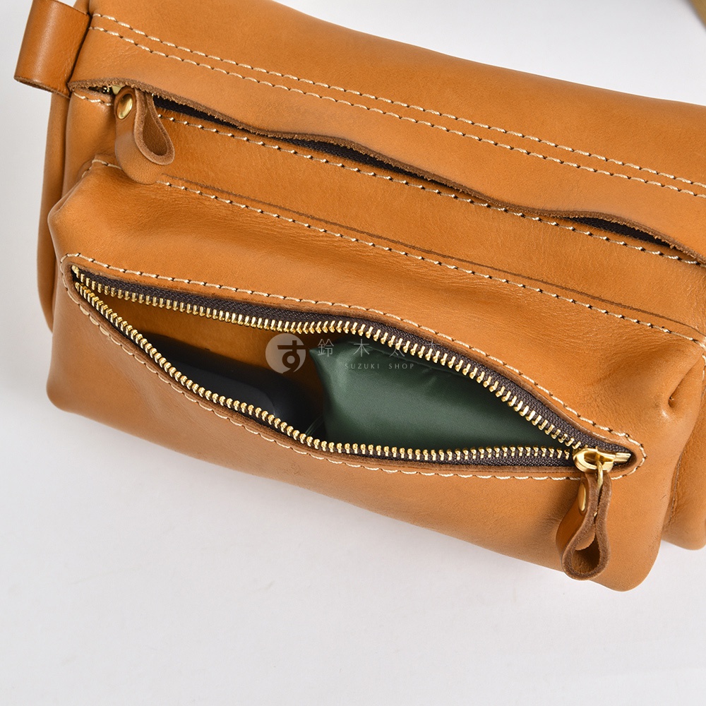 棕褐色皮革製成的手拿包，露出一個拉鍊開口的內袋，內有綠色物品，包身有縫線裝飾，金色拉鍊。