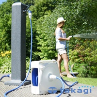 白色捲線器連接藍色水管，水管一端連接至灰色水龍頭柱，另一端由身穿條紋上衣與短褲、頭戴草帽的女性持握灑水槍澆灌花園。