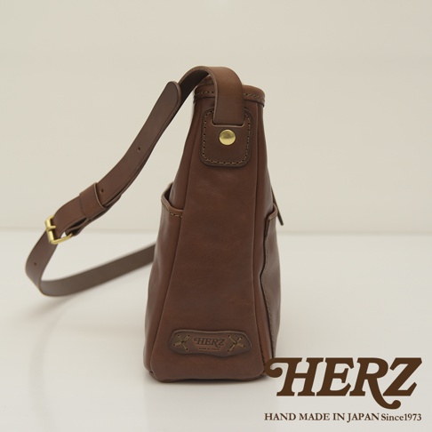 職人手作皮革輕便單肩側背包，採用棕色鞣革，設有可調節肩帶及金屬釦，底部有品牌標誌壓印。
