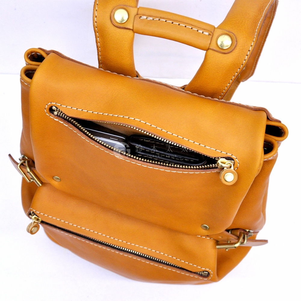 職人手作皮革多口袋後背包 R-135，採用溫暖的棕褐色皮革材質，設計有拉鍊開合的主袋與前袋，內部可見手機與皮夾等物品。黃銅色金屬配件與車縫線細節增添質感。