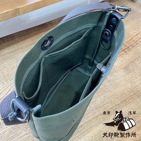 從頂部視角拍攝的綠色帆布斜背包內部，可見多個分隔口袋和磁釦，包帶為灰色織帶。圖片右下角有犬印鞄製作所的品牌標誌和日文文字。
