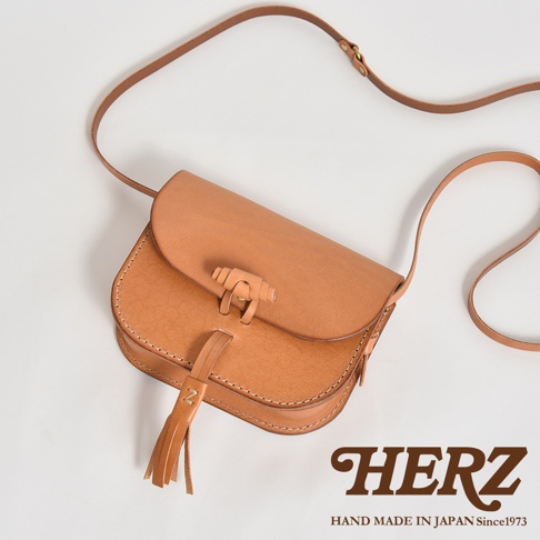 淺棕色皮革流蘇側背包，採用馬鞍包設計，配有可調節長度的細長皮革背帶和獨特的編織式鎖扣。包包正面有品牌標誌和流蘇裝飾。