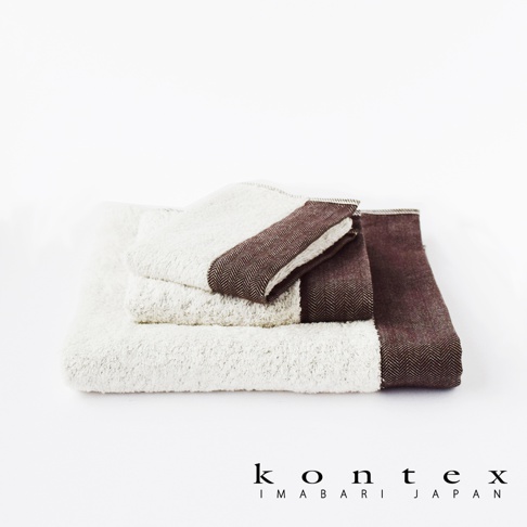 白色長纖有機棉毛巾堆疊，其中一條毛巾的邊緣飾有深棕色人字紋斜紋織帶。