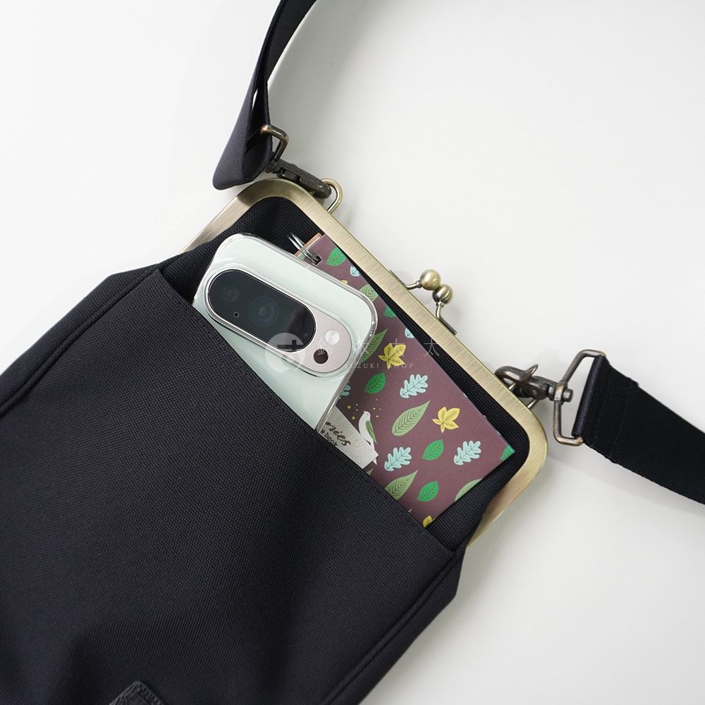 黑色斜背包，一個手機置於打開的古銅色口金包內，口金包內層印有樹葉圖案，包包有肩帶。