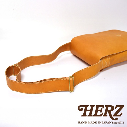 HERZ 品牌職人手作皮革軟皮直式斜背包，特寫鏡頭展現焦糖色皮革的細緻質感與縫線細節，搭配金屬釦件的肩帶。