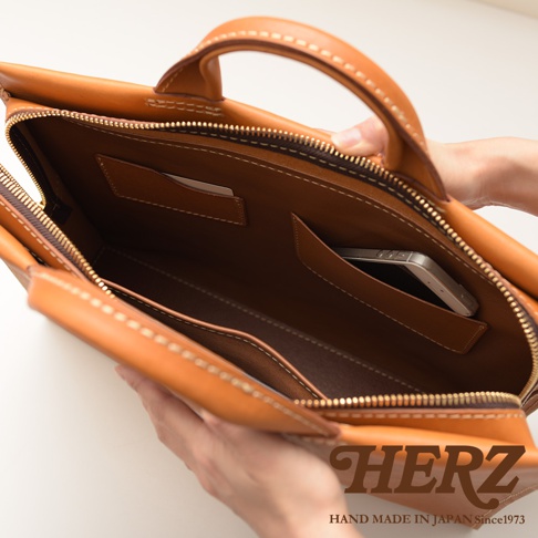 HERZ 品牌標誌，下方有 HAND MADE IN JAPAN Since1973 字樣，圖片為一個手工製作的皮革公事包內部，展示有手機和卡片插槽，以及縫線細節。