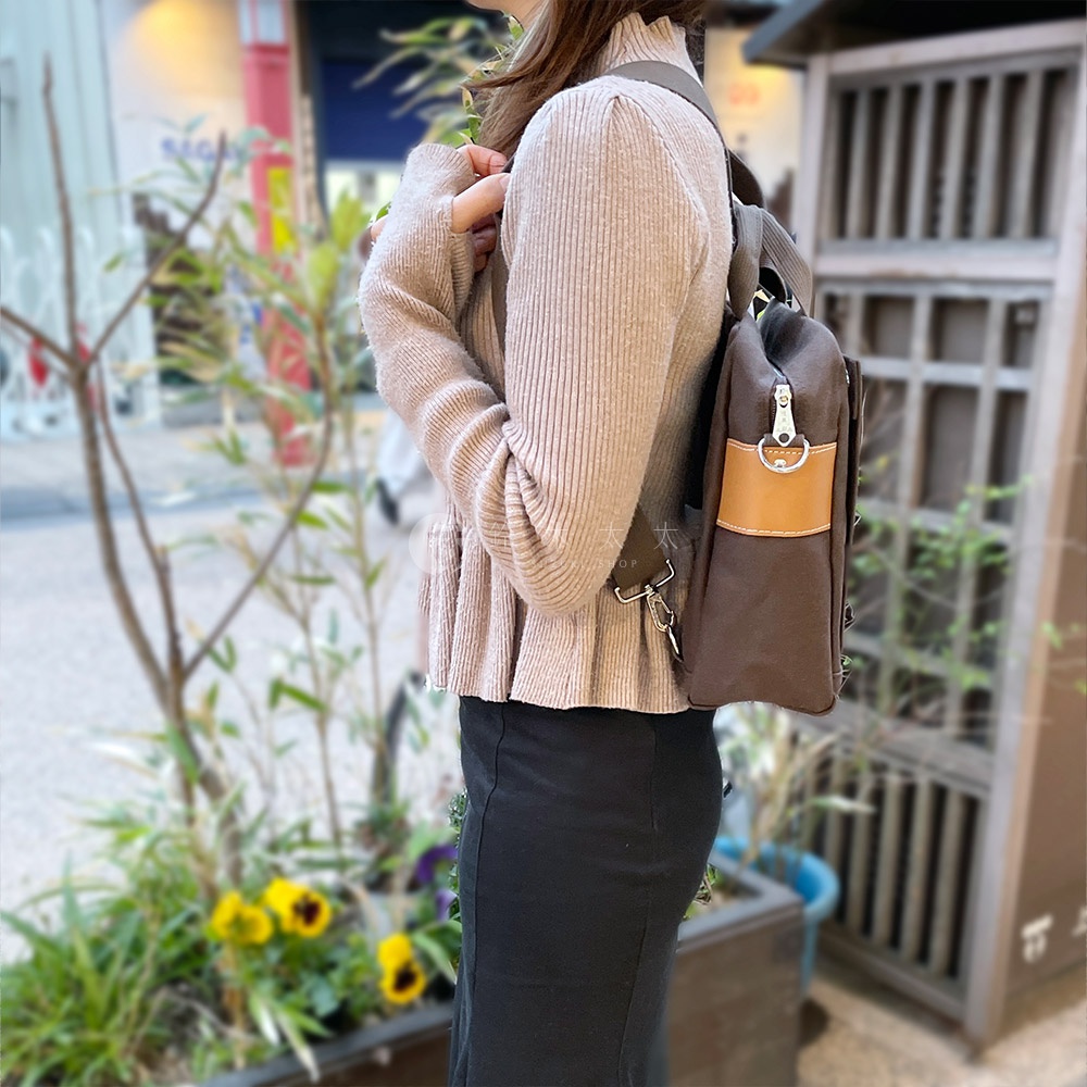 深棕色純棉帆布製成的三用橫式側背包，附有淺棕色皮革裝飾和銀色金屬配件，由一位女性模特兒以側背方式展示。