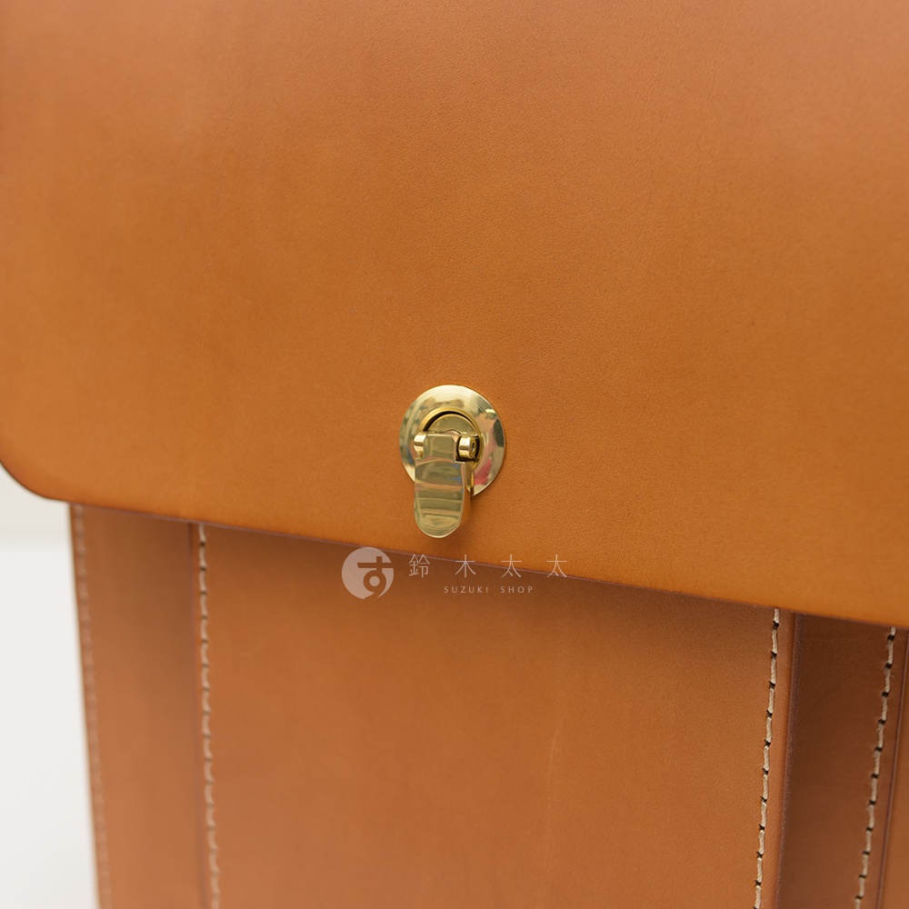 職人手作皮革復古後背包，以經典的棕褐色皮革製成，配有黃銅色旋轉鎖扣，側面有精緻的白色縫線。