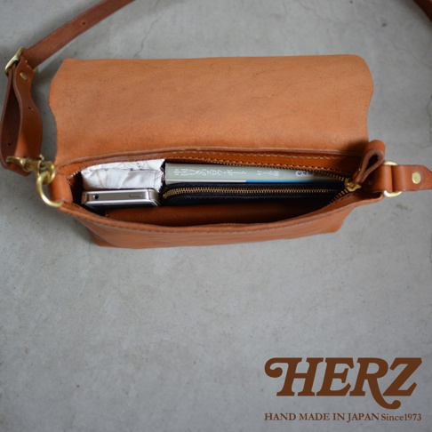 HERZ 品牌標誌，下方標示 HAND MADE IN JAPAN Since1973。一個棕色皮革斜背包，內部裝有手機、書本和錢包。