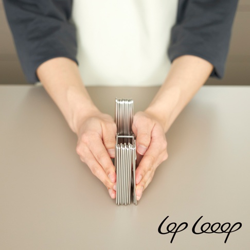 雙手正持握一個銀色不鏽鋼伸縮式瀝水架，架身由多個平行金屬桿組成，呈現折疊狀態，背景為淺灰色桌面及身著灰白色衣物的人。