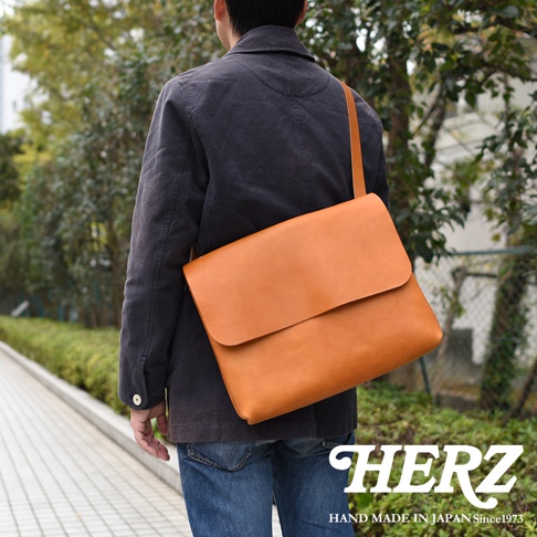 一位男士身著深色外套和牛仔褲，背著一個棕色皮革經典郵差包。包款設計簡約，有翻蓋和單肩帶。