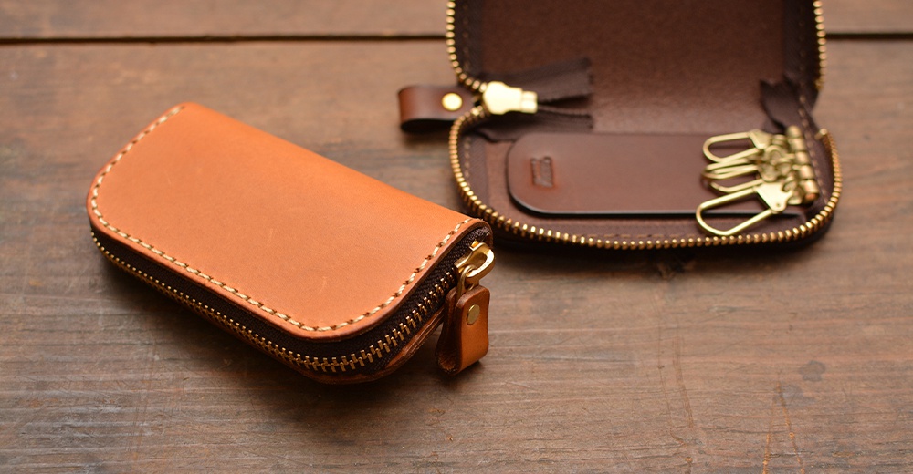 一個棕色皮革鑰匙包，上面有一個拉鍊，旁邊是打開的鑰匙包，露出四個金屬鉤。