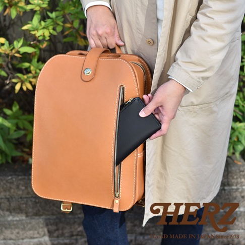 棕色皮革製作的兩用後背包，配有拉鍊前袋，一個黑色皮夾正被放入其中。包包邊緣有白色縫線裝飾，上方有提把和金屬扣。