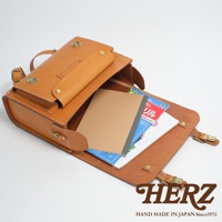 一個棕色皮革製成的橫式書包，包蓋打開，裡面放著幾本筆記本和書籍，包包的右下角有HERZ的商標。