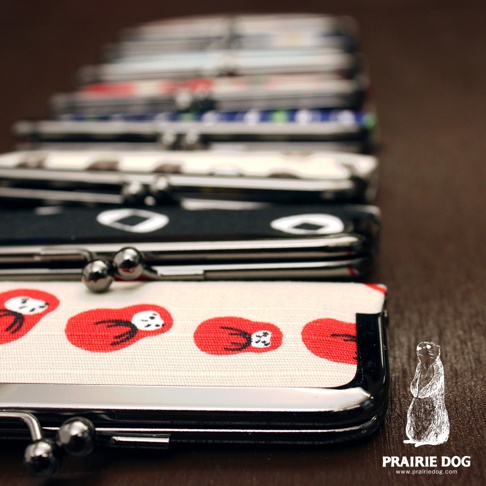 一排排和布華印鑑收納口金包，最前方為米白色布面印有紅色達摩圖案，搭配銀色金屬口金。後方有多款不同花色與圖案的口金包堆疊。