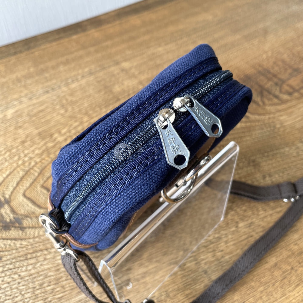 深藍色石洗帆布斜背手機包，具有雙層拉鍊設計，拉鍊頭上刻有「大印記」字樣。包包側面有棕色皮革飾邊和金屬掛環，搭配深棕色斜背帶。