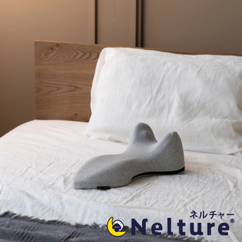 淺灰色布料製成的肩頸背拉伸牽引枕，呈現人體工學的彎曲造型，置於白色床單和木質床頭板旁。