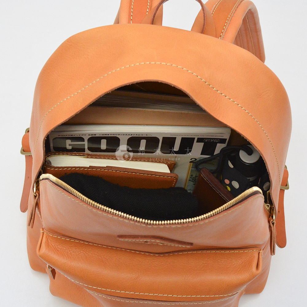 職人手作淺棕色皮革後背包，主隔層內有雜誌、筆記本、黑色毛衣、錢包，以及其他小物，展示其收納空間。