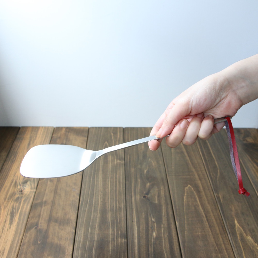 手持一把不鏽鋼萬用鍋鏟，鍋鏟呈銀色，鏟頭寬大呈彎月形。鍋鏟握柄處繫有一條紅色掛繩。