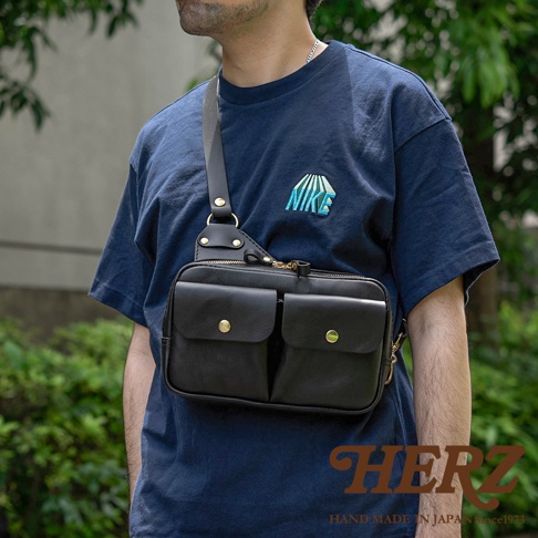 黑色皮革雙口袋斜背包，配有金色金屬釦和拉鍊，展示於身穿海軍藍 T 恤的男性身上。