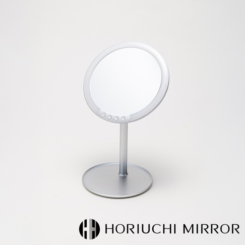 銀色金屬材質的桌上型化妝鏡，圓形鏡面周圍帶有LED燈，鏡面下方有四個圓形觸控按鈕。鏡子由一支細長的圓柱形支架連接至圓形底座。