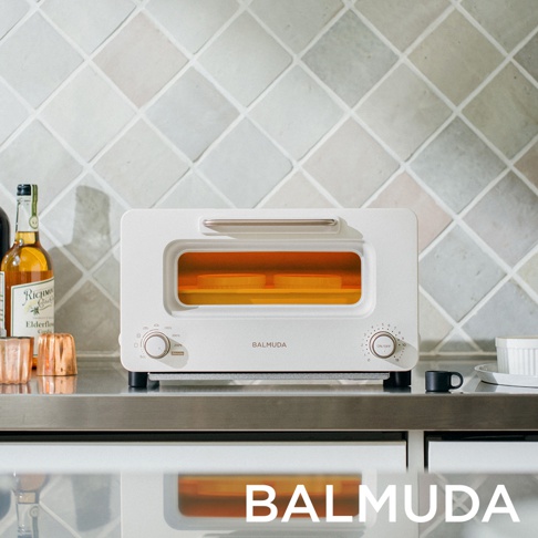 The white Balmuda The Toaster Pro K11C-SE sits on a stainless steel counter, with two slices of bread inside glowing orange.