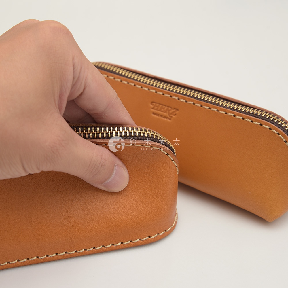 職人手作皮革三角弧形收納筆袋，採用自然棕色皮革，搭配金色拉鍊與米色縫線。其中一個筆袋被手握住，露出其精緻的材質與工藝。