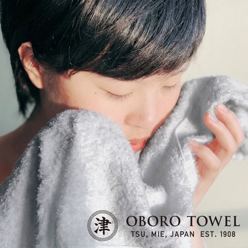 一位留著短髮的女性閉著雙眼，溫柔地將一條柔軟灰色的 Oboro Towel 品牌毛巾輕貼在臉頰上感受其細膩質感，圖片下方標示著 Oboro Towel 以及日本津市的創業年份 1908。
