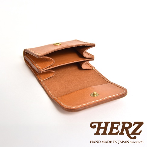 一個日本HERZ品牌職人手作的淺棕色皮革雙折零錢包，採皮革原色質感，正面呈現展開狀態，可見精緻的車縫線細節與金屬按扣，整體造型簡約俐落，下方印有HERZ品牌商標與產地資訊。