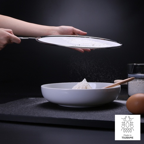 一只手拿著銀色金屬網篩，正在篩濾白色麵粉。麵粉篩入下方白色圓碗中，碗內已有部分麵粉。背景為黑色，畫面右側有雞蛋、木勺和裝有食材的透明罐。