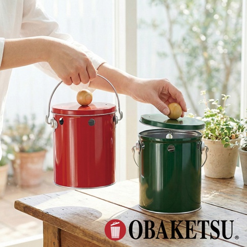 OBAKETSU 品牌標誌旁的日式手工製紅色和綠色蚊香桶，一隻手拿著紅色蚊香桶，另一隻手將綠色蚊香桶的木質圓球蓋子蓋上。