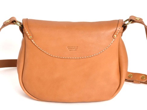 一款淺棕色皮革製成的馬鞍包，帶有翻蓋設計和縫線細節，配有可調節的皮革肩帶和金屬搭扣。