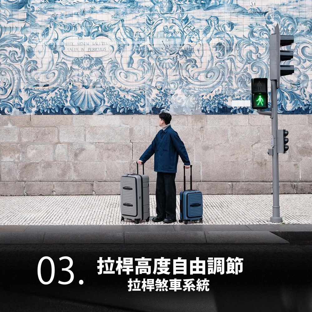 一位身穿深藍色外套與黑色長褲的男子站在人行道上，雙手分別拉著一個灰色與一個藍色的 AZPAC 前開口行李箱，背景為充滿藝術感的藍白配色葡萄牙瓷磚牆面，右側設有綠色行人號誌燈，畫面底部標註說明行李箱具備拉桿高度自由調節及拉桿煞車系統功能。