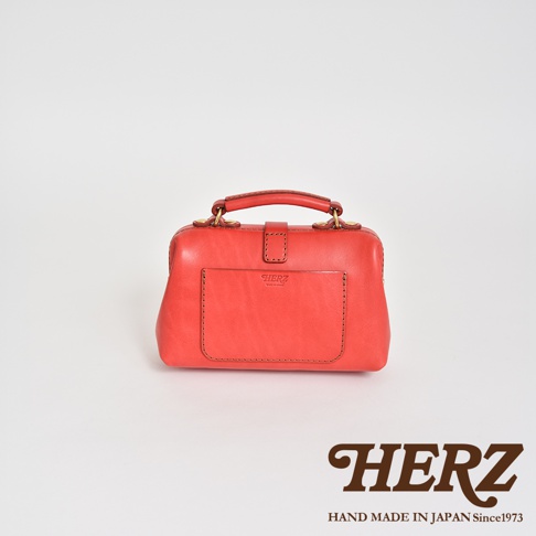 職人手作皮革迷你軟皮杜勒斯包，採用鮮豔的珊瑚紅色皮革製成，配有金色金屬扣和提把。包包正面有一個貼袋，上面壓印有品牌標誌。