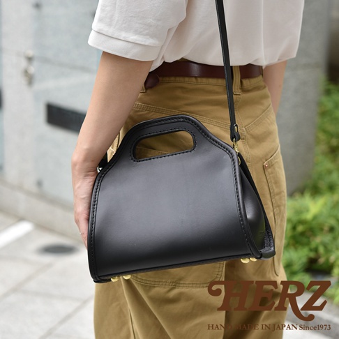 黑色皮革製作的職人手作單肩兩用拱型手袋，配有手提握把和可調節的長肩帶。包身線條流暢，頂部有縫線細節，底部設有金屬腳釘。