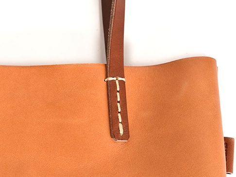 職人手作皮革直式軟皮托特包，採用光滑的駝色皮革，搭配咖啡色皮革提把，以白色縫線加固。