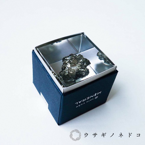 深藍色方形盒內，一個銀色金屬立方體容器中，置放著一塊具有金屬光澤、多面結晶結構的黃鐵礦礦石。