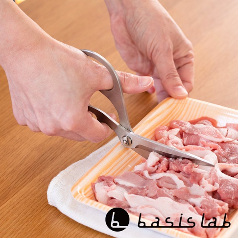 一雙銀色不鏽鋼萬用料理剪刀，左手正握住剪刀，右手扶著裝有生肉的橘色塑膠盒，剪刀的刀刃正在切割肉塊。