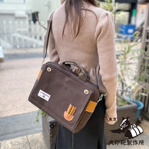 棕色純棉帆布製成的橫式側背包，配有皮革飾邊和提把，可肩背或手提。