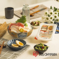 一碗裝有生魚片、芥末和蔥花的日式丼飯，旁邊有兩個溏心蛋、一小碟毛豆、一瓶水，以及一包標示「低溫製法微波白飯 150g」的產品。