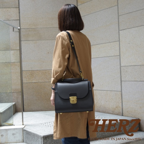 一位女子身著駝色長袍，斜背著一個黑色的皮革手提包，包包上有一個金色的鎖扣。包包下方有HERZ品牌的標誌和「HAND MADE IN JAPAN Since1973」的文字。