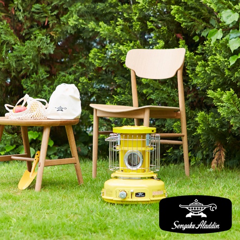 黃色卡式瓦斯暖爐，設有圓形爐頭和金屬防護網，底部有控制旋鈕和品牌標誌，放置在綠色草地上，背景為樹叢和木質椅子。