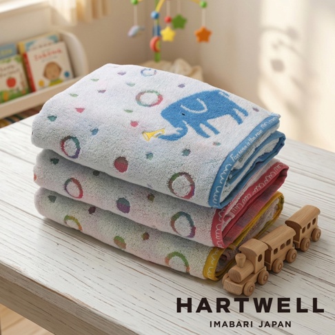 一張疊放的 Hartwell 今治雨中遊行小象浴巾，白色毛巾上印有可愛藍色小象與彩色圈圈圖案，邊緣帶有彩色縫線細節，旁邊擺放著一個木製火車玩具，背景為溫馨的嬰兒房擺設。