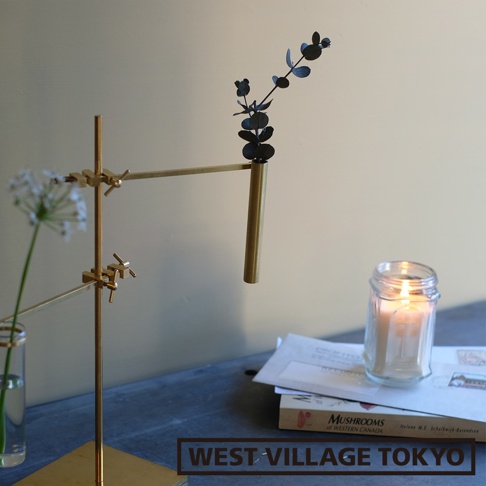 黃銅材質的支架上，夾著一個細長的黃銅花筒，花筒內插著一株黑色枝葉植物。背景為淺灰色牆面，桌面上有一玻璃瓶裝白色小花、一本雜誌和一罐點燃的蠟燭。