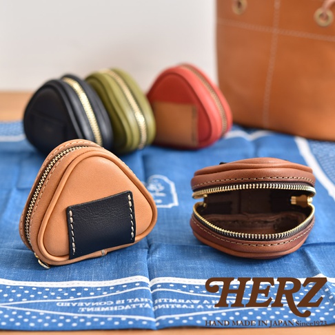 數個職人手作皮革飯糰造型小物收納包，包含棕色、黑色、綠色和紅色。其中一個棕色收納包特寫，拉鍊半開，露出內部。