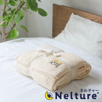 米白色絨毛毯，表面有細緻的條紋紋理，以白色緞帶繫綁，並附有標有「Cheese MOFU」的吊牌。毯子疊放在白色床單上，背景為木製床頭板和綠色植物。